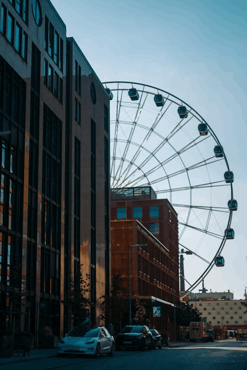 „Blick durch eine urbane Straße im Münchner Werksviertel mit modernem Riesenrad im Hintergrund – umgeben von Bürofassaden, Autos und bunten Gebäuden, aufgenommen bei Tageslicht mit klarem Himmel.“