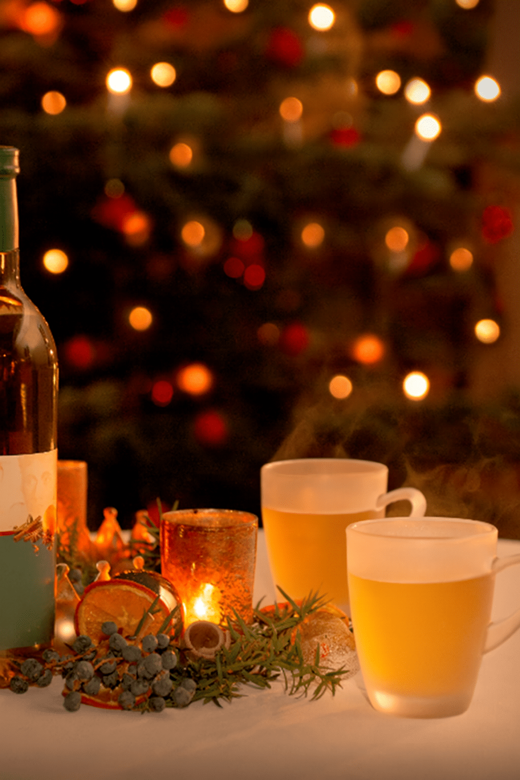 Festlich dekorierter Weihnachtstisch mit dampfenden Glühweintassen der Marke „Wackerbarths Weiß & Heiß“, Flasche mit Gewürzwein, leuchtenden Kerzen, Tannenzweigen und getrockneten Orangen vor einem stimmungsvoll beleuchteten Weihnachtsbaum – ideal für exklusive Weihnachtsfeiern, Winterevents und kulinarischen Genuss in festlicher Atmosphäre.