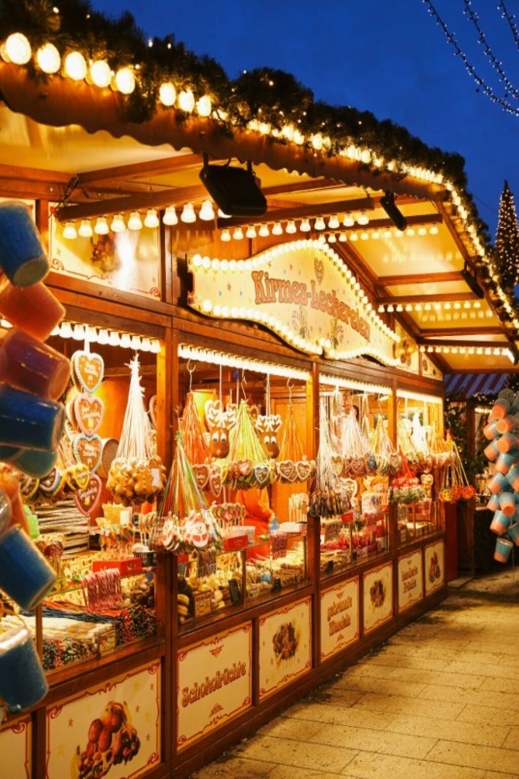 Festlich beleuchteter Weihnachtsmarktstand mit traditionellen Süßwaren wie Lebkuchenherzen, Zuckerstangen und gebrannten Mandeln. Die weihnachtlich geschmückte Bude versprüht nostalgischen Charme und lädt zum Genießen und Verweilen ein – ideal für stimmungsvolle Teamevents und Weihnachtsfeiern in der Adventszeit.