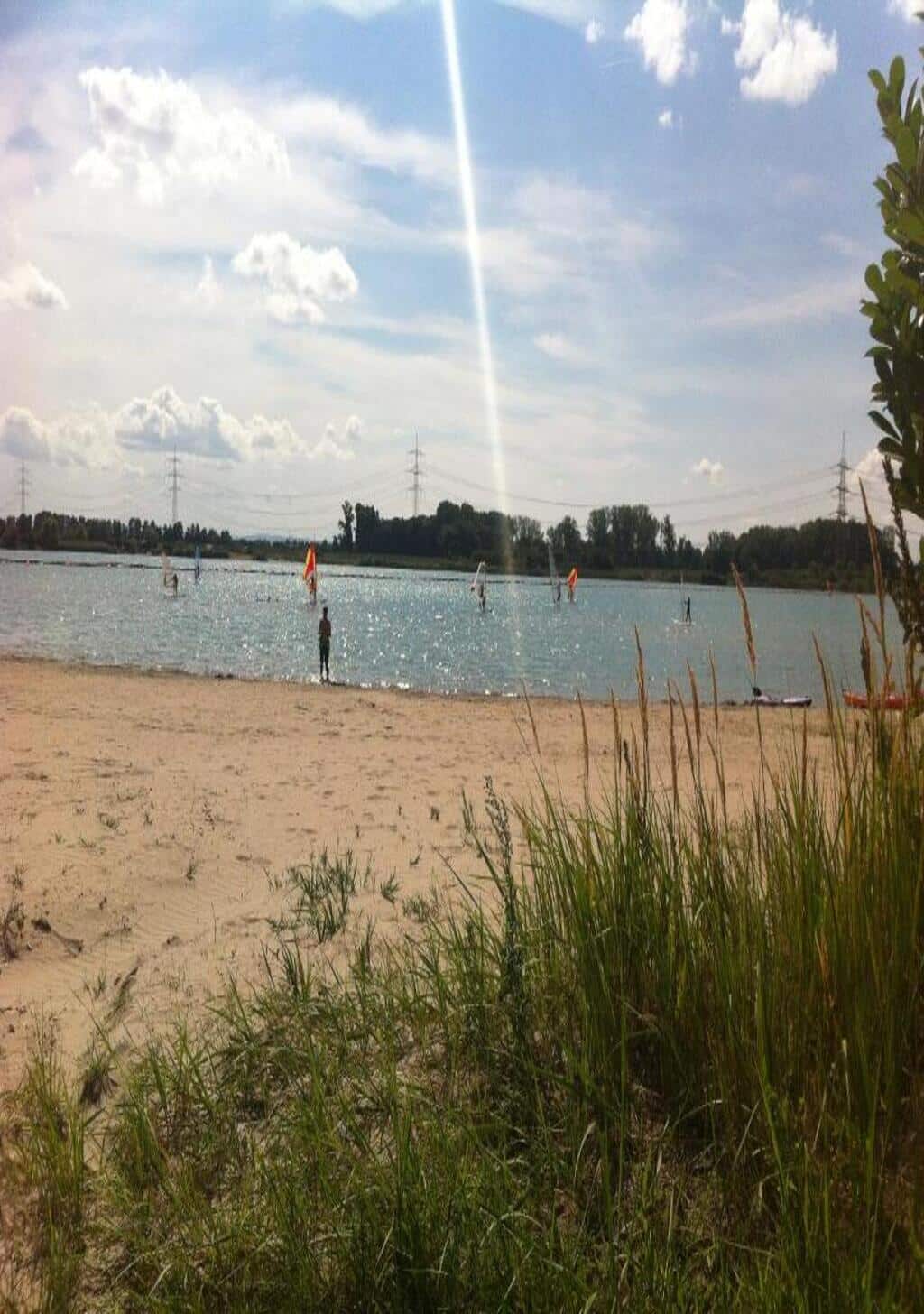 Sandstrand und Blick auf den Riedsee mit Wassersportlern – idyllische Kulisse für ein Teambuilding mit Beachfeeling in entspannter Sommeratmosphäre.